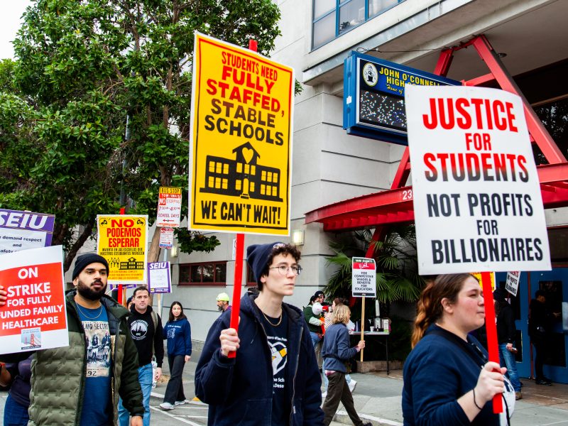 A group of people participate in a protest, carrying signs demanding well-staffed schools, student justice, and fully funded healthcare.