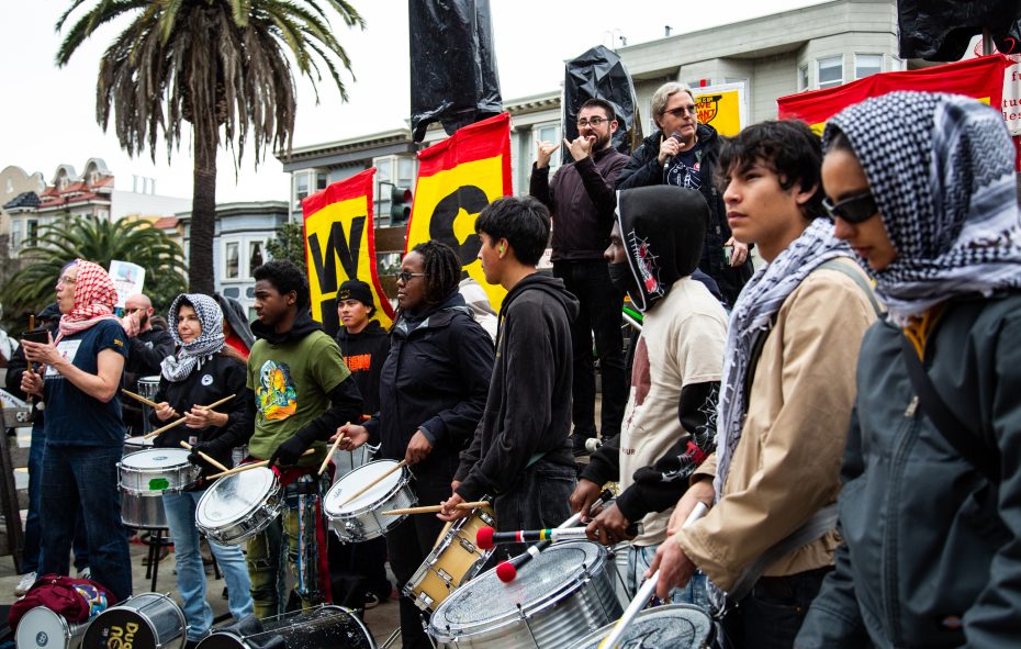 A group of people stand outdoors with drums and banners, some wearing scarves over their heads, participating in a protest or demonstration.