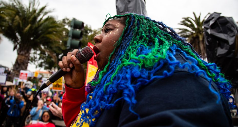 A person with long blue and green hair speaks into a microphone at an outdoor event, with a crowd and palm trees visible in the background.