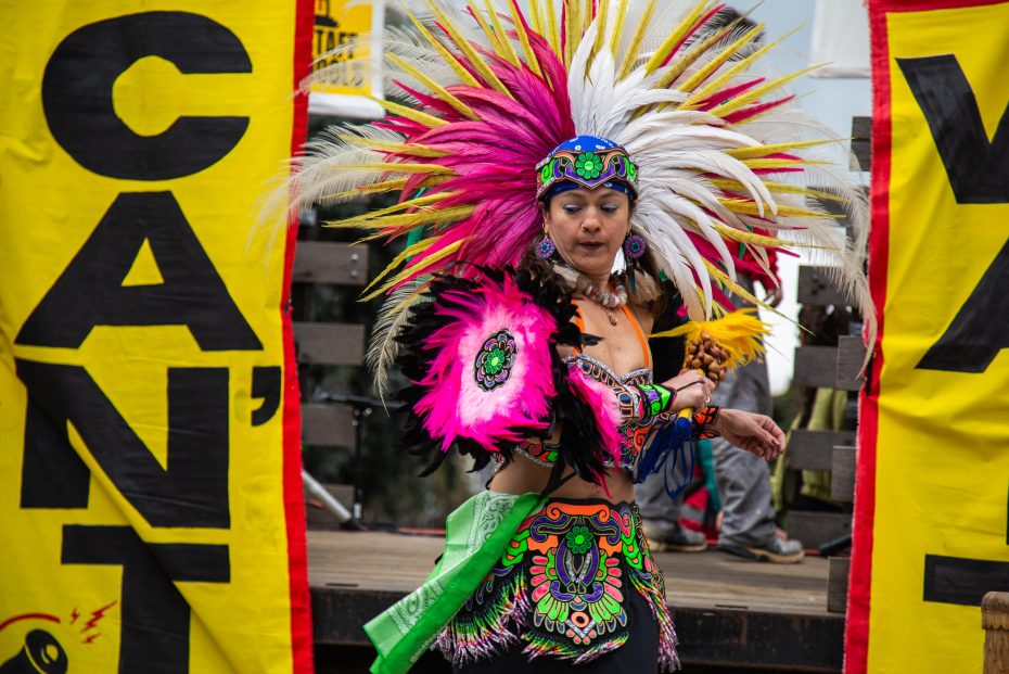 A dancer in vibrant traditional attire and feathered headdress performs in front of yellow banners with bold black text.
