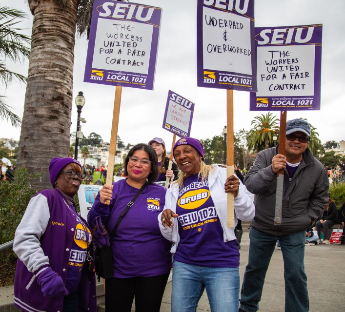 Four people wearing SEIU shirts and holding protest signs advocating for fair contracts stand together outdoors, smiling at the camera.