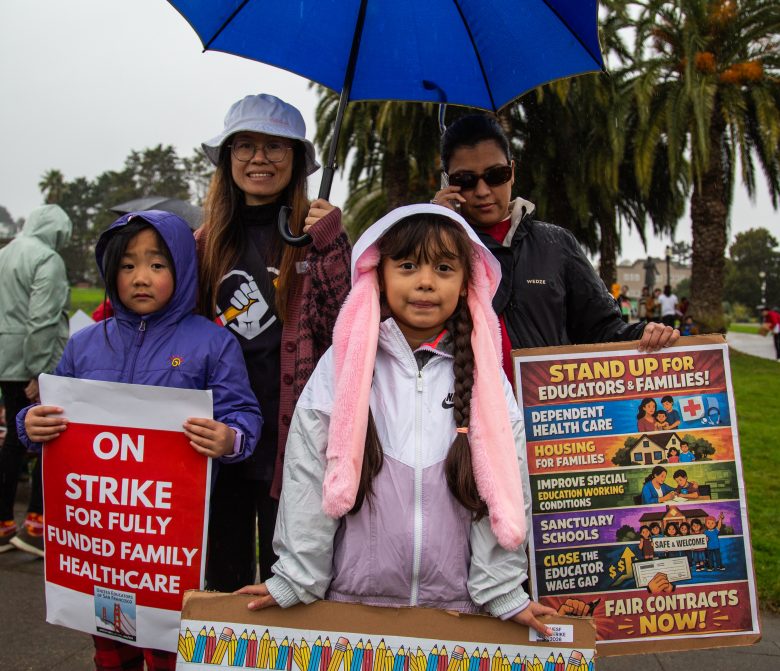 A group of adults and children stand outside in rain gear at a protest, holding signs advocating for fully funded family healthcare and fair contracts for educators.