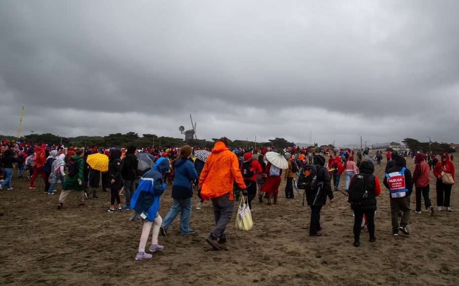 A large crowd of people, many in raincoats and carrying umbrellas, walk on a cloudy, wet beach.