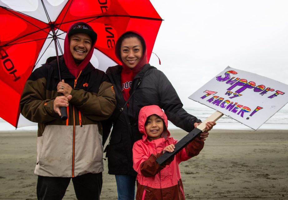 A smiling family stands on a beach under an umbrella. The child holds a handmade sign that reads, "Support my teacher!.