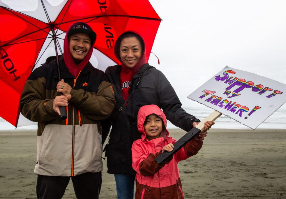Three people stand on a beach under a red and white umbrella; a child holds a sign that reads “Support my teacher!” in colorful letters.