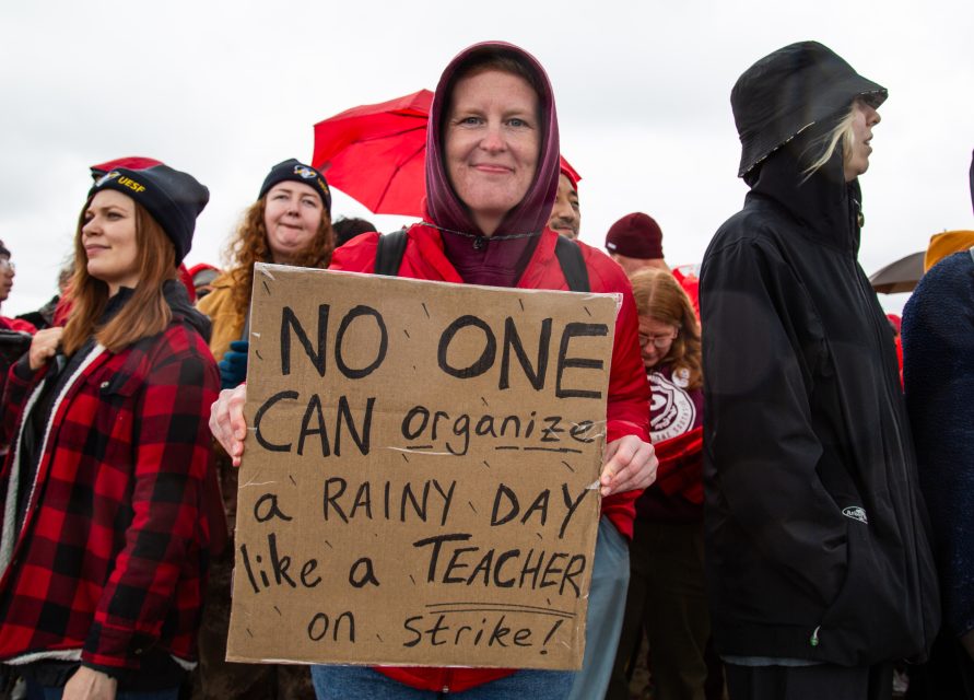 A person holds a sign reading "NO ONE CAN organize a RAINY DAY like a TEACHER on strike!" while standing in a crowd on a rainy day.
