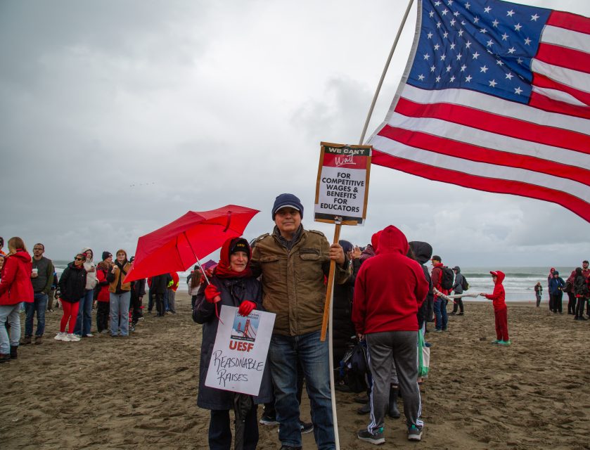 Two people stand on a beach holding protest signs and a large American flag, surrounded by a crowd under cloudy skies.
