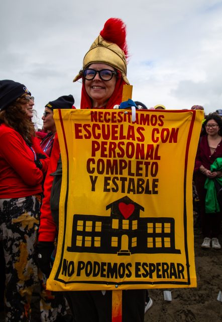 Person wearing a helmet holds a yellow sign in Spanish reading, "We need schools with full and stable staff. We can't wait," during a public gathering.