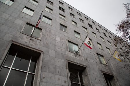 Low-angle view of a gray stone building with multiple windows and three flags, including the U.S. and California flags, hanging outside.