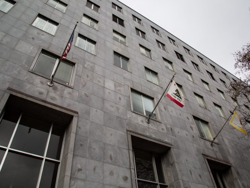 Low-angle view of a gray stone building with multiple windows and three flags, including the U.S. and California flags, hanging outside.