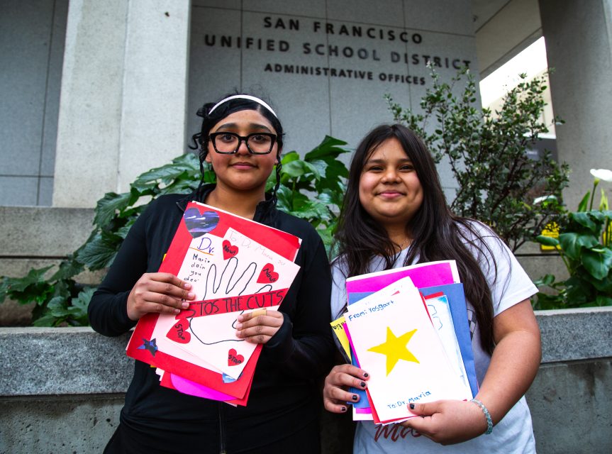 Two students stand in front of the San Francisco Unified School District building, each holding handmade signs and cards.