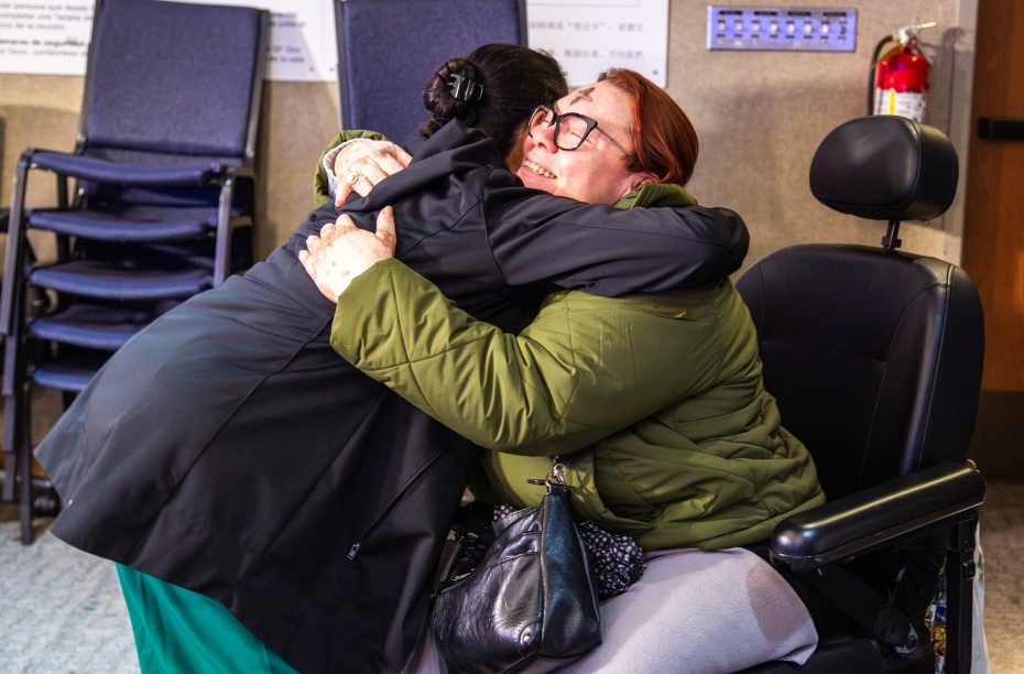 Two women hug each other warmly; one is sitting in a wheelchair and smiling, while the other stands and embraces her. A stack of chairs and a fire extinguisher are visible in the background.