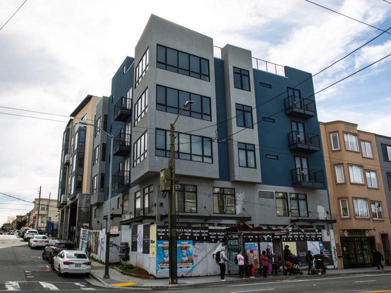 A modern four-story apartment building on a street corner with people gathered at the entrance and cars parked along the curb.