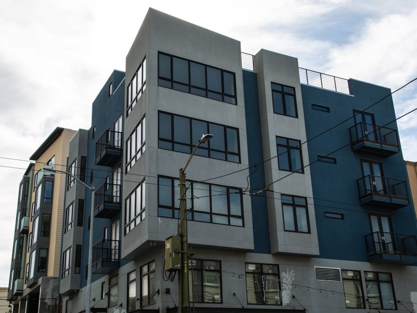 Modern four-story apartment building with blue and gray exterior, large windows, and balconies, under a partly cloudy sky.