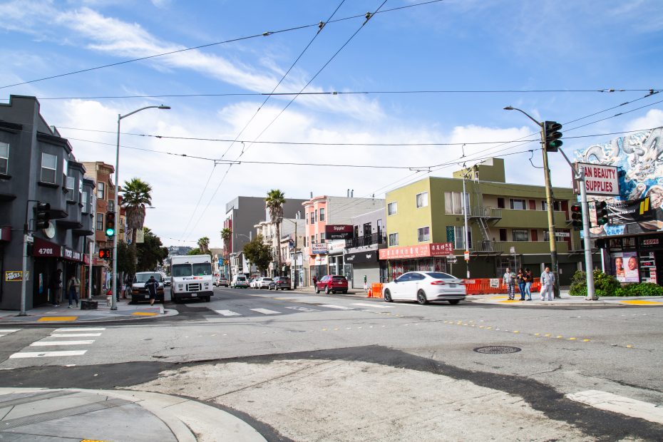 A city street intersection with cars, pedestrians, shops, and buildings under a partly cloudy sky. Traffic lights and overhead wires are visible.