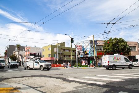 Intersection with traffic, utility and service vehicles, crosswalks, overhead wires, and buildings with murals and storefronts under a partly cloudy sky.