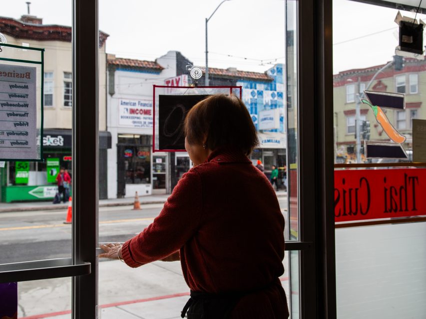 A person in a red sweater stands inside by a glass door, looking out onto a city street with shops and signs visible across the road.