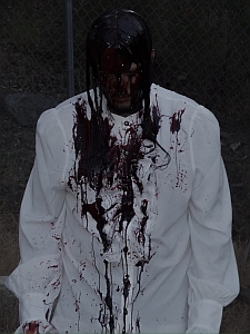 A person in a white shirt is covered in what appears to be dark, blood-like liquid, standing outdoors with a fence in the background.