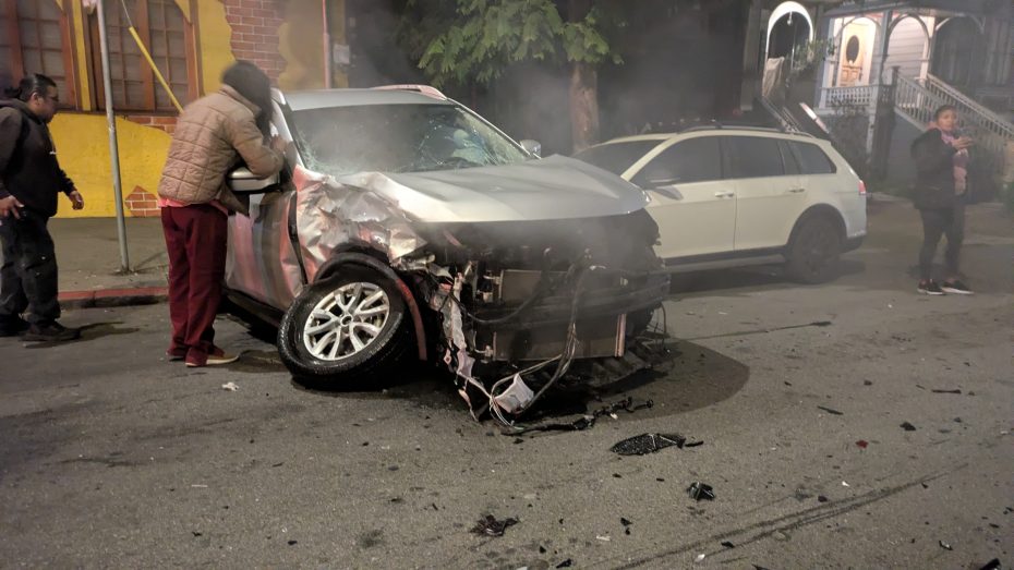 A heavily damaged silver SUV with a crumpled front is parked on a street at night; a person stands beside it while debris is scattered on the ground.