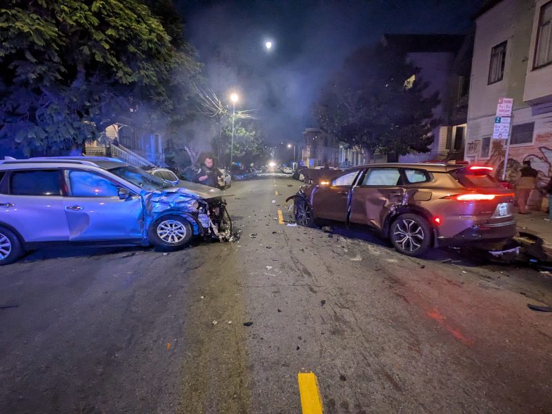 Two cars with significant front-end damage are stopped in the middle of a city street at night, debris scattered, with people and a dog standing nearby.