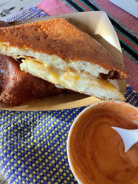 Two halves of a grilled cheese sandwich in a cardboard tray next to a small cup of orange dipping sauce with a plastic spoon.
