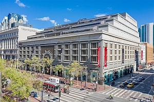 A large multi-story department store building with red banners on a busy city street corner, with cars, pedestrians, and a clear blue sky.