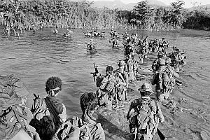 Group of soldiers in full gear wade through a river in a jungle environment, carrying rifles and supplies. Trees and mountains are visible in the background.