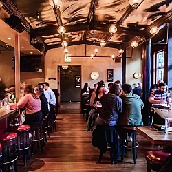 People are dining and socializing at a cozy, dimly lit restaurant with a bar on the left and tables along the right side.