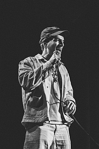 A person wearing a cap and casual clothes holds a microphone and speaks on stage. The image is in black and white.