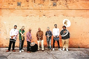 Seven musicians stand in a row against an orange brick wall, each holding their respective brass or percussion instruments.