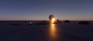 A metallic, spiked structure stands on a flat surface at sunset, with a person and several objects nearby under a clear sky.