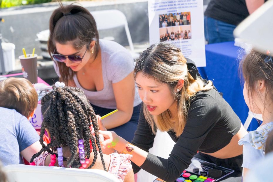 A woman paints a girl's face at an outdoor event while others watch and participate around a table with art supplies.