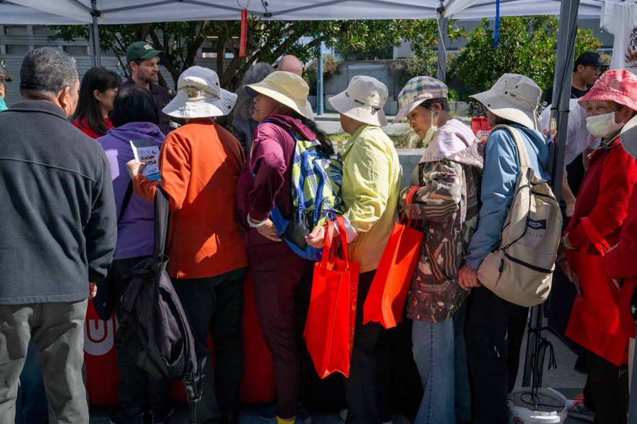 A group of people wearing hats and backpacks stand in line under a canopy, holding red tote bags at an outdoor event.