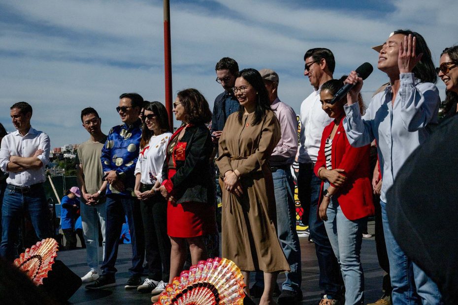 A group of people stand on an outdoor stage, some smiling and one person speaking into a microphone, with decorative fans on the ground in front of them.