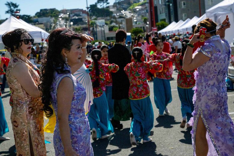 Women in ornate dresses stand in the foreground while a group in bright costumes perform a choreographed dance at an outdoor street festival with white tents.
