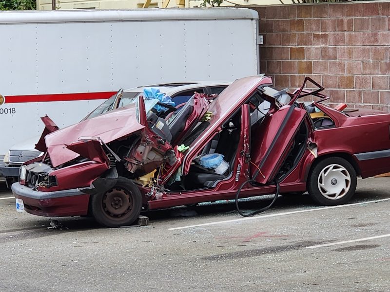 A severely damaged maroon sedan with deployed airbags parked in front of a white truck and a brick wall. The car is crushed from the front and side, exposing the interior.