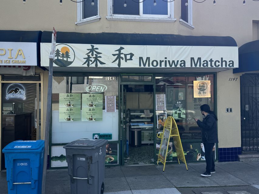 Storefront of Moriwa Matcha with a person standing outside, an open sign, a yellow sandwich board, and two blue trash bins on the sidewalk.