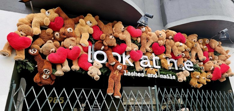 Dozens of teddy bears holding red hearts are arranged above the entrance of a store called "look at me lashes and nails." A metal gate is partially visible in front.