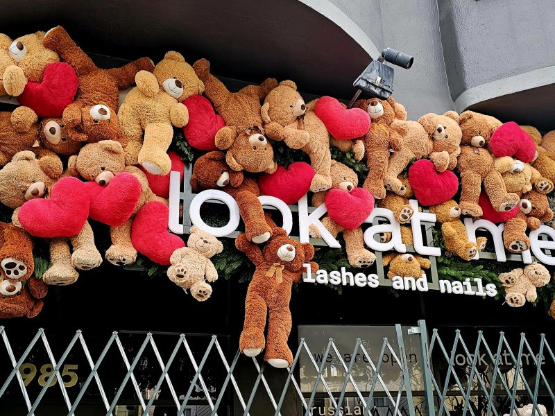 Dozens of teddy bears holding red hearts are arranged above the entrance of a store called "look at me lashes and nails." A metal gate is partially visible in front.