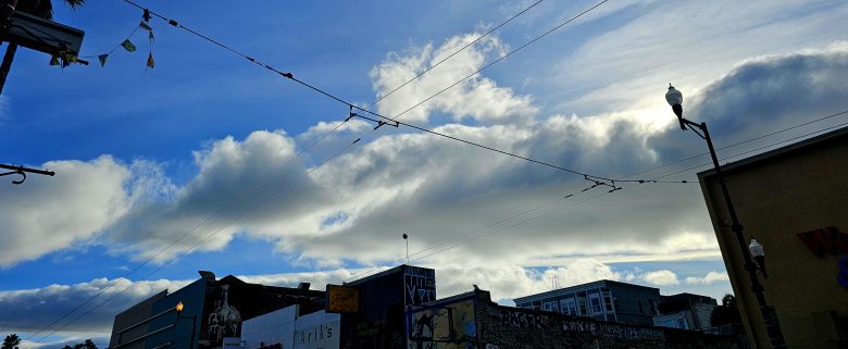 Overhead view of a city street with clouds in the sky, utility wires crossing, streetlights, and building rooftops visible below.