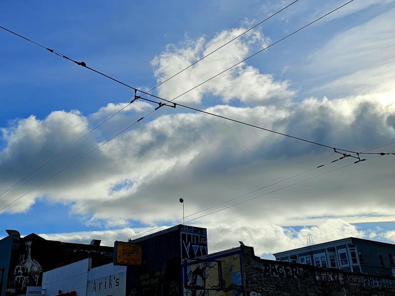 Overhead view of a city street with clouds in the sky, utility wires crossing, streetlights, and building rooftops visible below.
