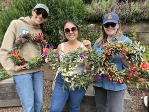 Three women standing outdoors hold handmade wreaths decorated with colorful flowers and greenery. They are smiling and wearing sunglasses and casual clothing.