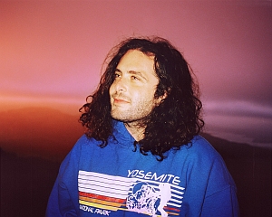 A person with long curly hair wearing a blue "Yosemite National Park" hoodie stands outdoors at sunset, looking slightly upward.