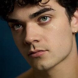 A young person with curly dark hair and blue eyes looks slightly off camera with a neutral expression, a single tear visible on their cheek, against a dark background.