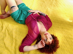 Person with curly hair lies on a yellow quilt, wearing a magenta top and green shorts. One arm is raised, and tattoos are visible on their arms and thigh.
