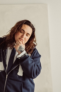 Person with long brown hair wearing a navy jacket and white shirt stands in front of a plain background, resting their chin on their hand and looking downward.