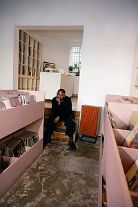 A person dressed in black sits on a step between shelves of records in a modern, well-lit room with a concrete floor.