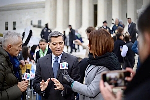 A man in a suit speaks to reporters holding microphones in front of a large government building with columns, as people gather in the background.