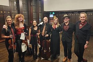 Seven people, some holding string instruments, stand together indoors, smiling at the camera. They are dressed in semi-formal and performance attire.