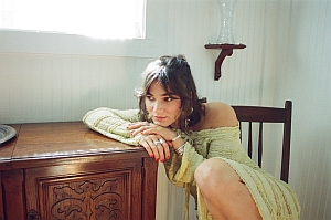 A woman in a light green off-the-shoulder dress sits on a wooden chair, leaning on a wooden cabinet in a sunlit room.